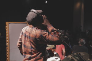 Filmmaker Yemi Oyediran holds on to his headset with his right hand while standing in front of a microphone, recording music for Queen City Kings