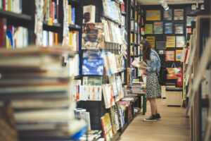 A woman reads a book while surrounded by other books in an aisle of a bookstore